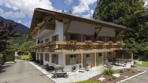 A two-story chalet with wooden balconies, a patio area with outdoor seating, and a mountain backdrop. - Apartment in Grainau am Badersee (Graifenau)
