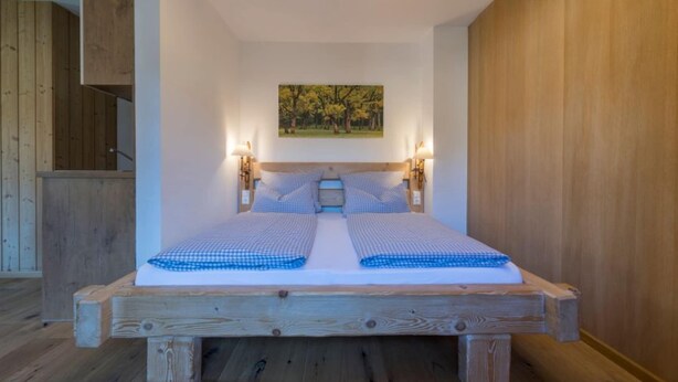 A wooden bed with a checkered bedspread, two bedside lamps, and a painting on the wall. - Apartment in Grainau am Badersee (Graifenau)