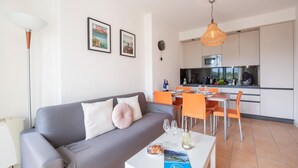 A modern living room with a grey sofa, a dining table with orange chairs, and a kitchen in the background. - Appartement in Porlezza am Luganersee (Porlezza)