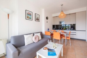 A modern living room with a grey sofa, a dining table with orange chairs, and a kitchen in the background. - Appartement in Porlezza am Luganersee (Porlezza)