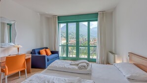A hotel room with two beds, a desk, and a chair. There is a view of mountains and a city through the window. - Appartement in Porlezza am Luganersee (Porlezza)