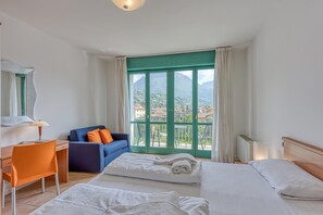 A hotel room with two beds, a desk, and a chair. There is a view of mountains and a city through the window. - Appartement in Porlezza am Luganersee (Porlezza)