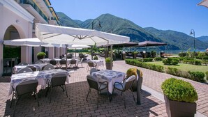 An outdoor dining area with tables and chairs, umbrellas, and a view of mountains and water. - Appartement in Porlezza am Luganersee (Porlezza)
