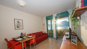 A compact living space with a dining area, a red sofa, and a view of the outdoors. - Appartement mit Seeblick auf den Luganersee (Porlezza)