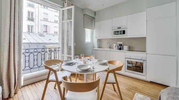 Een moderne keuken met witte kasten, een magnetron en een oven. Er staat een eettafel gedekt met borden, glazen en een kaars. De kamer heeft houten vloeren en uitzicht op een gebouw door een raam met een metalen hekwerk.
