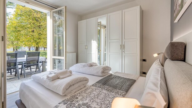 A bedroom with a bed, a bedside table, a chair, and a view of an outdoor area with a table and chairs. - Appartement mit Seeblick auf den Luganersee (Porlezza)