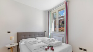 A neatly made bed with white linens and pillows, a bedside table with a lamp, and a window with curtains. - Appartement mit Seeblick auf den Luganersee (Porlezza)