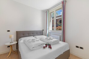 A neatly made bed with white linens and pillows, a bedside table with a lamp, and a window with curtains. - Appartement mit Seeblick auf den Luganersee (Porlezza)