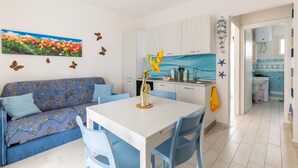 Un soggiorno moderno con divano, tavolo da pranzo e sedie. C'è un angolo cottura con mobili bianchi e uno schienale blu. Un quadro con fiori adorna la parete, e sono presenti decorazioni a forma di farfalle e stelle marine.