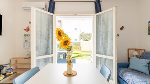 Una stanza con un tavolo bianco, sedie blu, un letto con biancheria a motivi e vista su un giardino attraverso una porta vetrata.