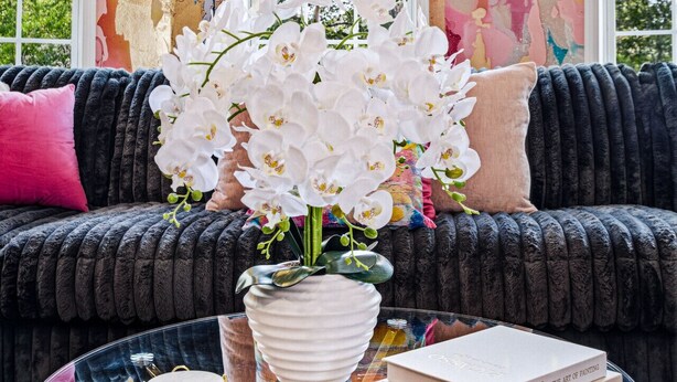 A living room with a colorful abstract wall, a black sofa with pink and beige pillows, a glass coffee table with a white vase and books, and a window with a view of trees.