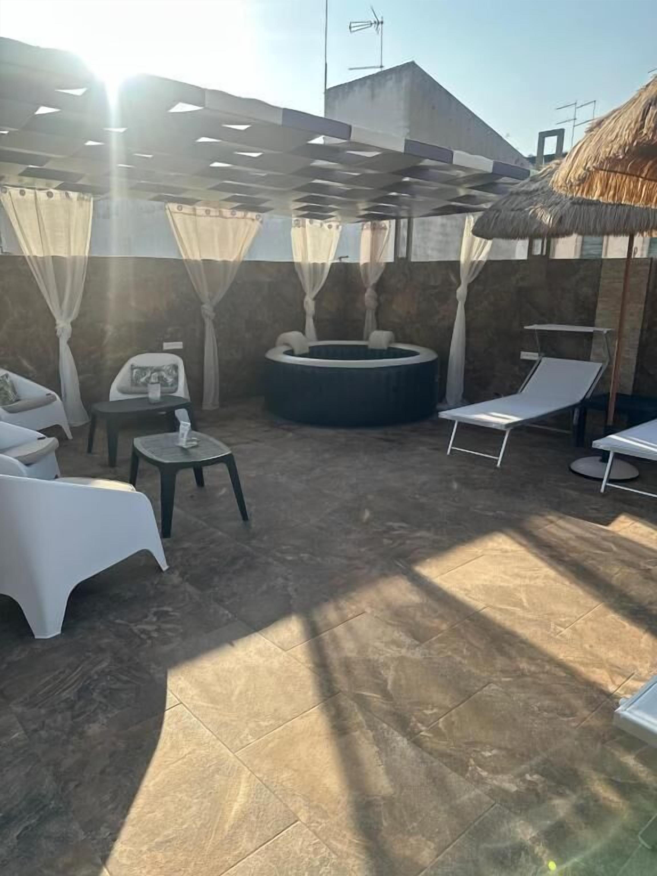 A sunlit outdoor patio with a hot tub, lounge chairs, and a small table.
