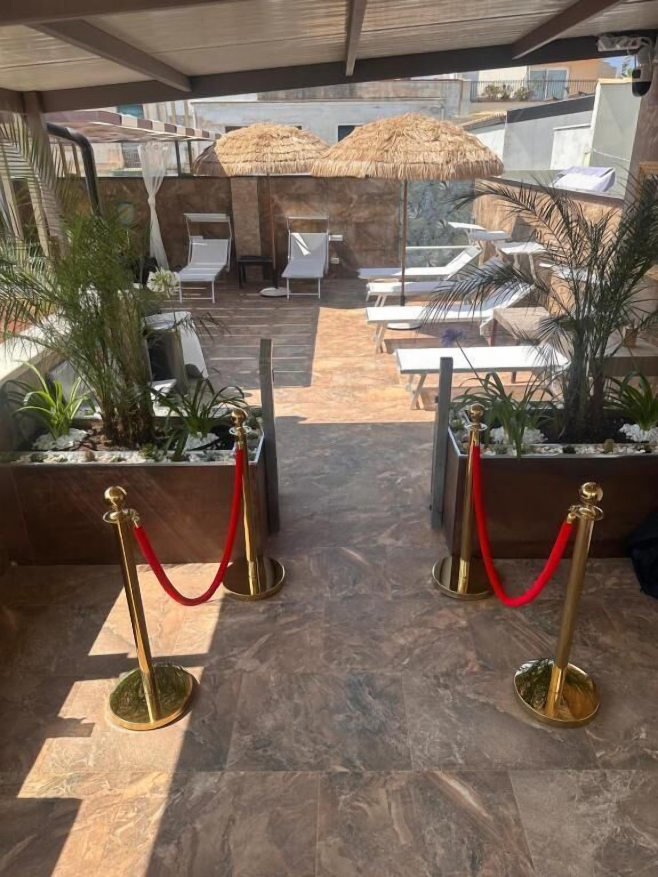 Un área de lounge al aire libre, iluminada por el sol, con sillones blancos, sombrillas de paja y cuerdas de terciopelo rojo que delimitan una sección privada.
