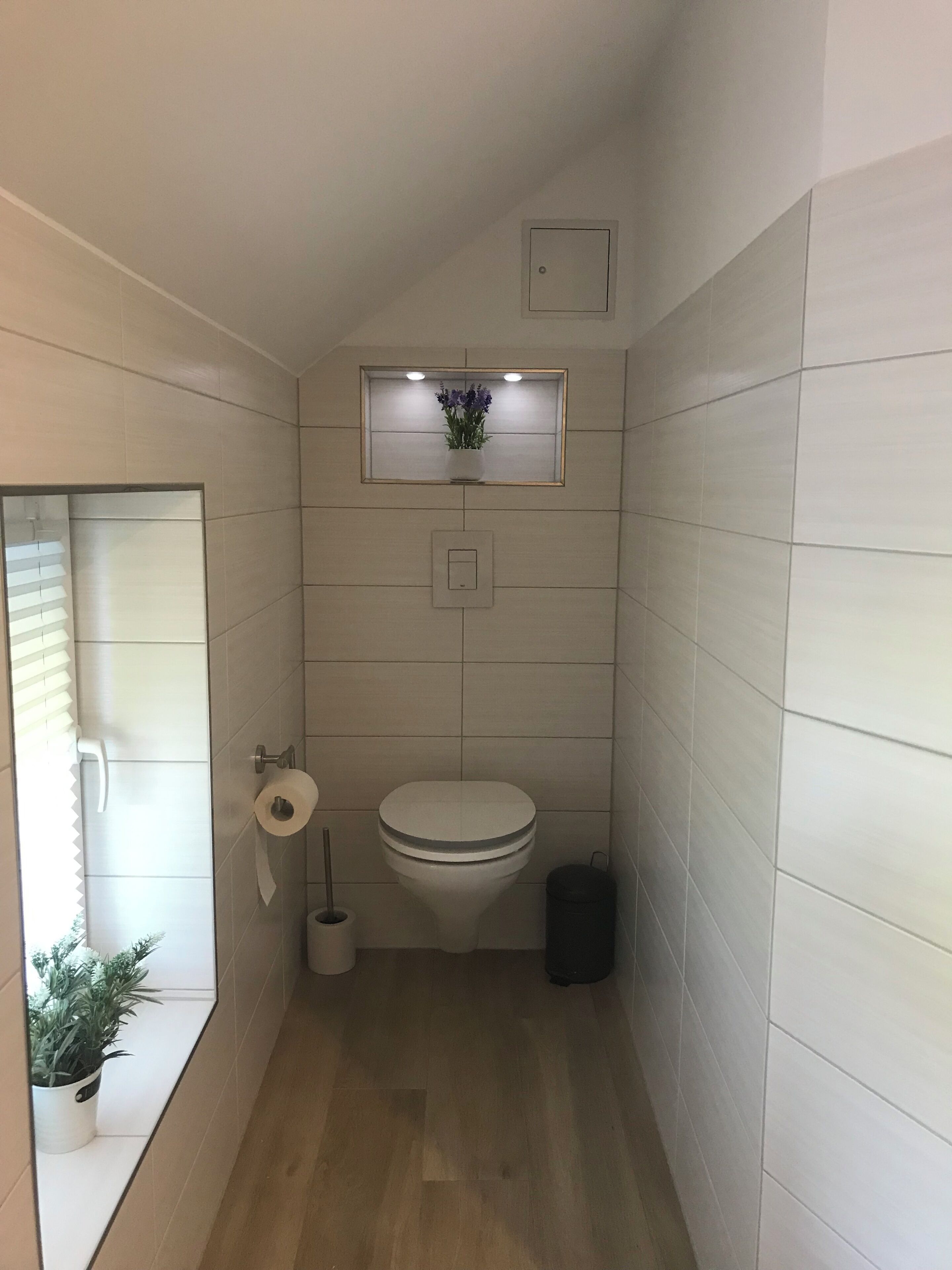 A small, modern bathroom with a toilet, a wall-mounted shelf with a plant, a toilet paper holder, and a trash bin.