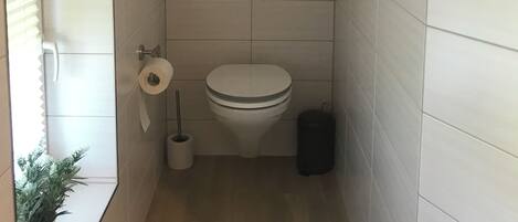 A small, modern bathroom with a toilet, a wall-mounted shelf with a plant, a toilet paper holder, and a trash bin.