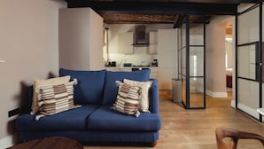 A modern living room with a blue sofa, two pillows, and a wooden coffee table. A kitchen is visible in the background with white cabinets and a stainless steel range hood. - FiveM High Street Kensington (London)