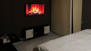 Een slaapkamer met een bed, een aan de muur bevestigde televisie waarop een haard te zien is, een kledingkast en een klein tafeltje met spullen erop.