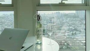 Ein Laptop auf einem Tisch mit einer Flasche und einem Weinglas, und Blick auf eine Stadtlandschaft durch große Fenster.