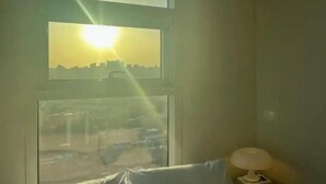 Ein Zimmer mit großem Fenster, einer Lampe auf einem Nachttisch und Blick auf eine Skyline bei Sonnenuntergang.