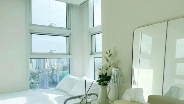 Ein modernes Hotelzimmer mit einem großen Bett, einem Nachttisch mit Lampe, einer Vase mit Blumen und Blick auf die Stadt durch große Fenster.