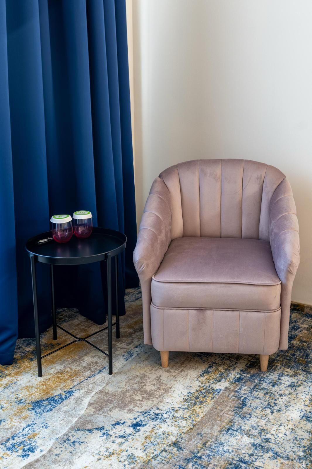 Una habitación de esquina con un sillón color crema, una mesita redonda con dos botellas y una cortina azul.