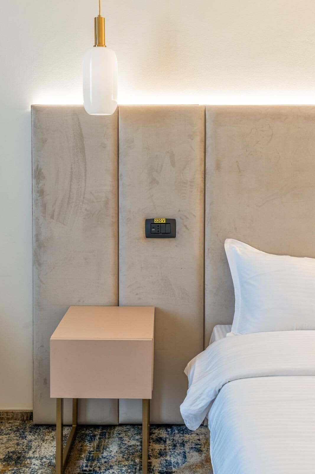 Quarto de hotel com cama, mesa de cabeceira, tomada na parede.