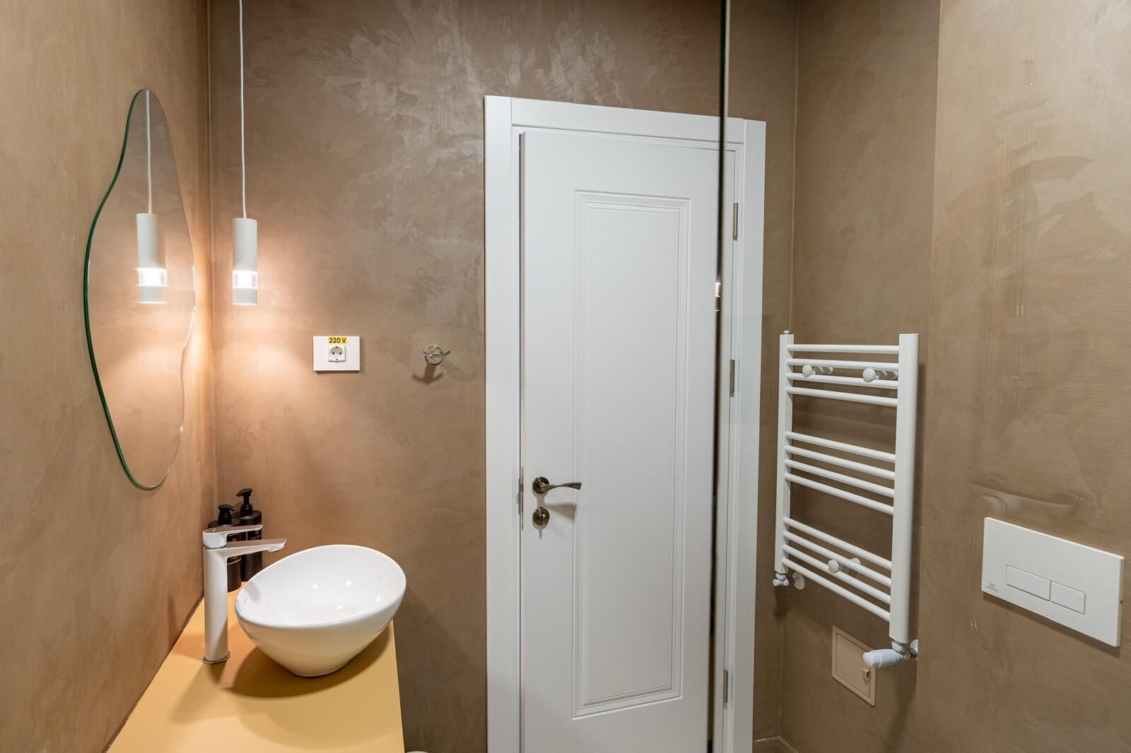 Un baño con puerta blanca, lavamanos blanco, toallero y ducha con puerta de cristal.