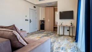 A modern hotel room with a beige sofa, a flat-screen TV mounted on the wall, a small table with a vase and a bottle, and a wardrobe with a radio. - Hotel Sierra (Pitesti)