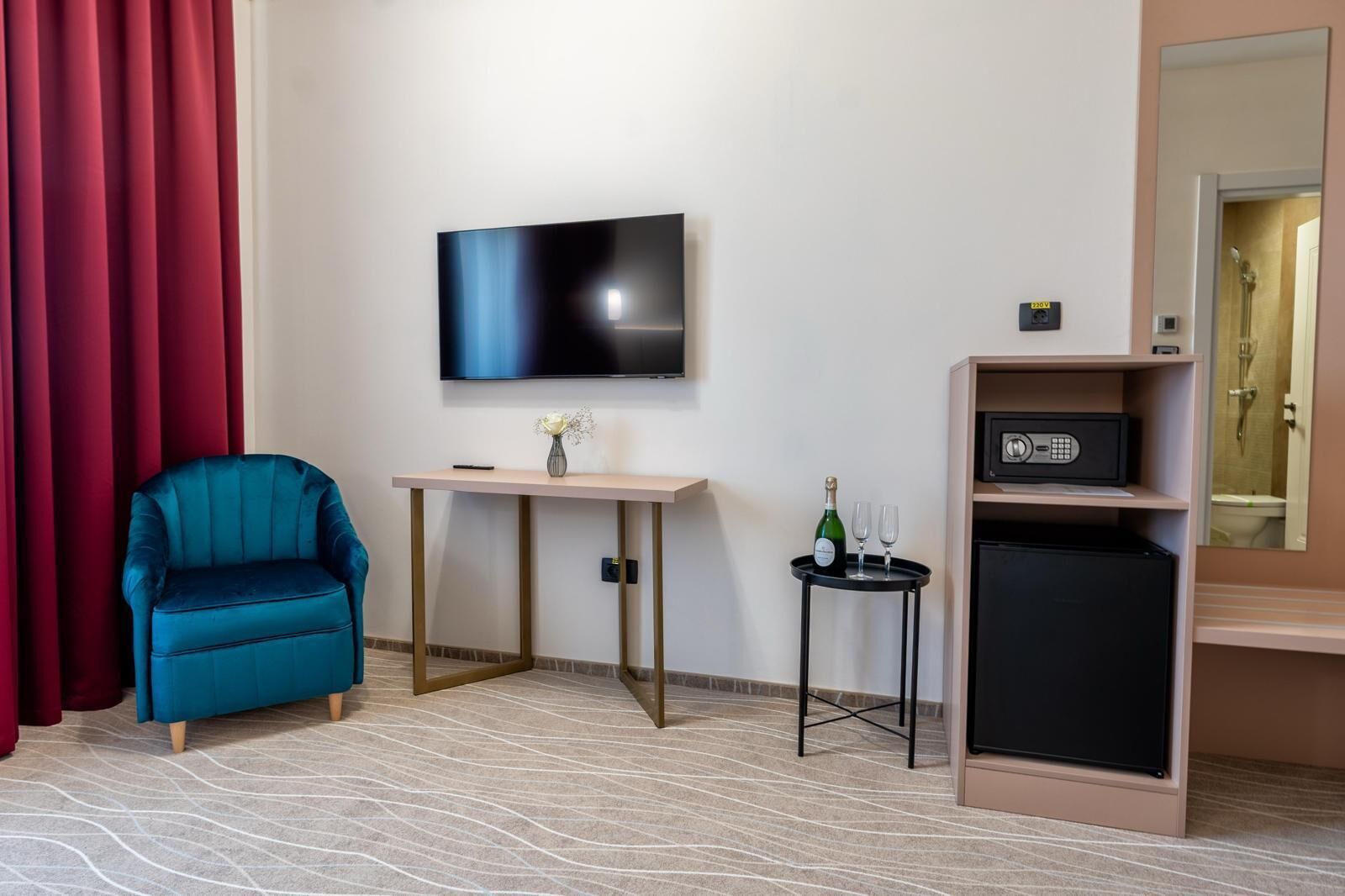 A hotel room with a TV, a small table with a vase and a bottle of champagne, a blue armchair, and a minibar.