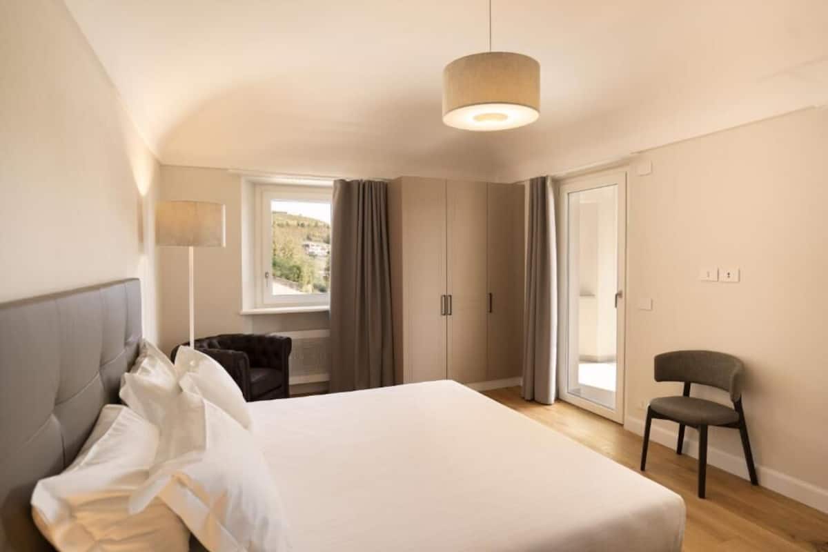 A bedroom with a large bed, a chair, a wardrobe, and a window with curtains.