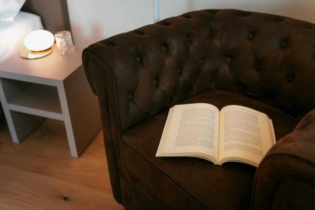 Un fauteuil marron à motifs capitonnés, avec un livre ouvert posé dessus, une table de chevet avec une lampe et un plancher en bois.