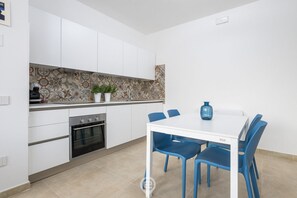 A modern kitchen with white cabinets, a tiled backsplash, and a dining area with a white table and blue chairs. - Coru e Mari Appartamenti - Baia (Pula)