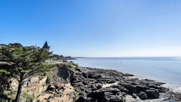 A coastal landscape with rocky terrain, a clear sky, and a distant building with a pointed roof.