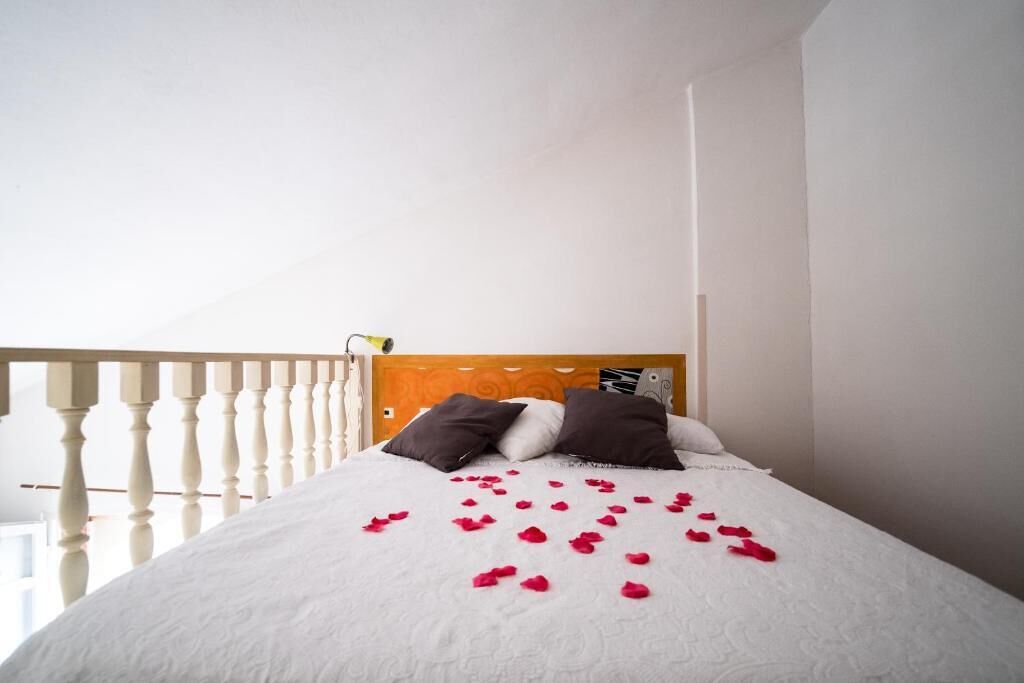 A neatly made bed with white bedding and brown pillows, a wooden headboard, and red rose petals scattered on the bed.