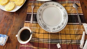 Een eettafel met diverse gerechten, waaronder ananasschijfjes, kokoskoek en een bord met wat lijkt op een dessert. Op de tafel staan ook een waterfles, een kop koffie en een smartphone.