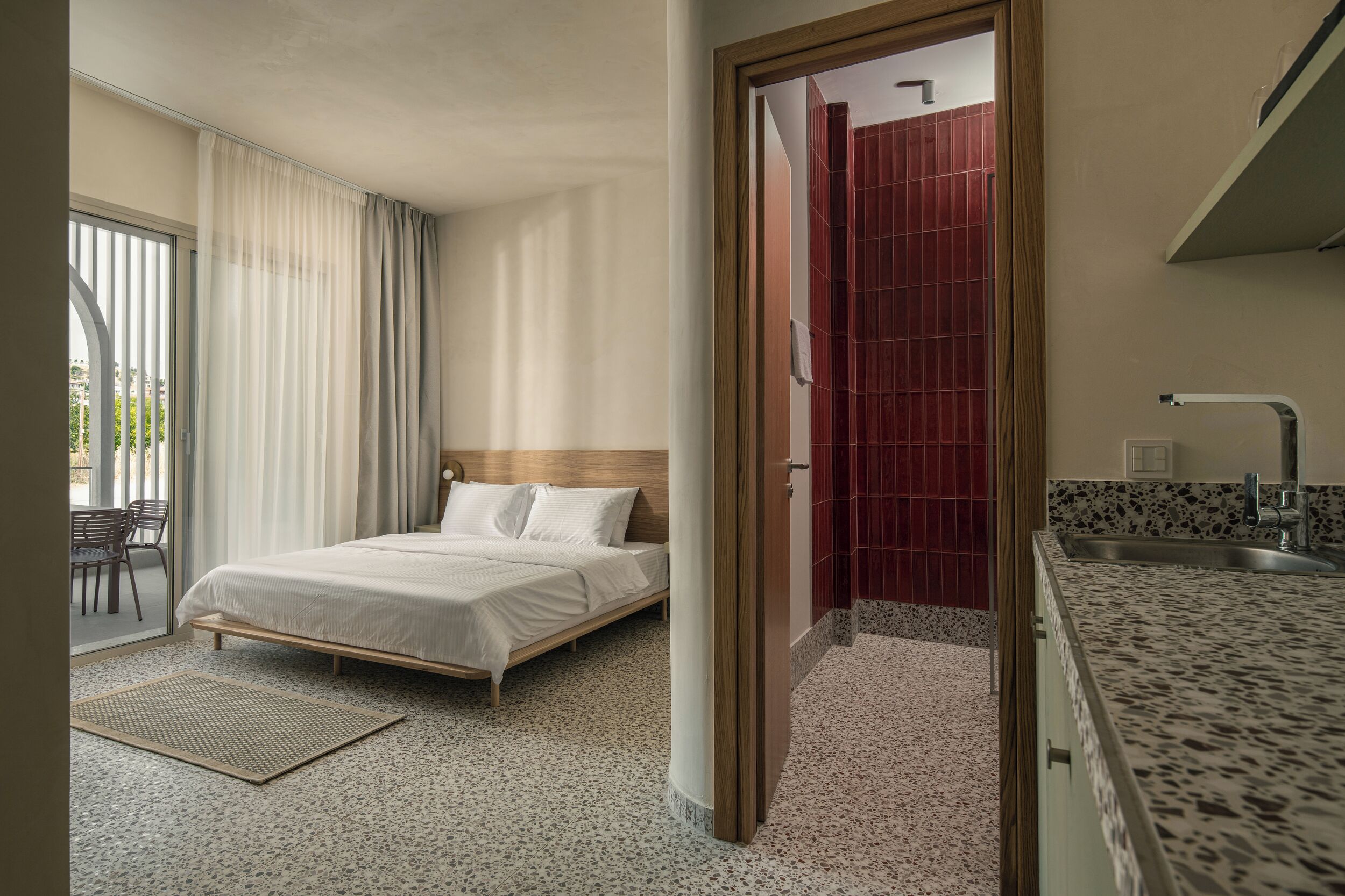 A hotel room with a bed, a small sitting area with a chair, a kitchenette with a sink, and a view through a door to a red-tiled bathroom.