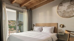 A hotel room with a large bed, a wooden headboard, a bedside table with a lamp, and a view of the city through the balcony. - FOS Luxury Suites (Nikiti)