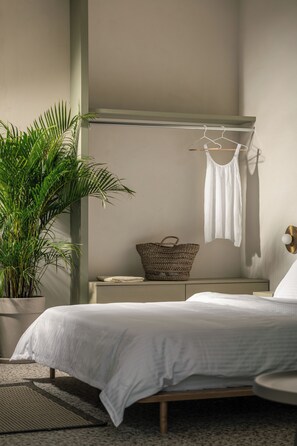 Un dormitorio con una cama, un tendedero, una planta en maceta, una cesta tejida y una mesita de noche.