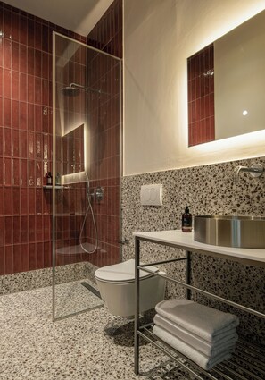 Un baño moderno con cabina de ducha de vidrio, inodoro blanco, lavabo con pileta de acero inoxidable y toallero con toallas dobladas.