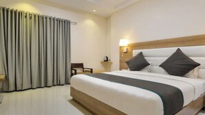 A hotel room with a large bed, a wooden headboard, a nightstand, a chair, and a window with curtains. - Hotel Blue Orchid (Udaipur)