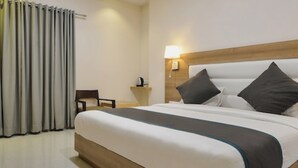 A hotel room with a large bed, a bedside table with a lamp, a chair, and a window with curtains. - Hotel Blue Orchid (Udaipur)