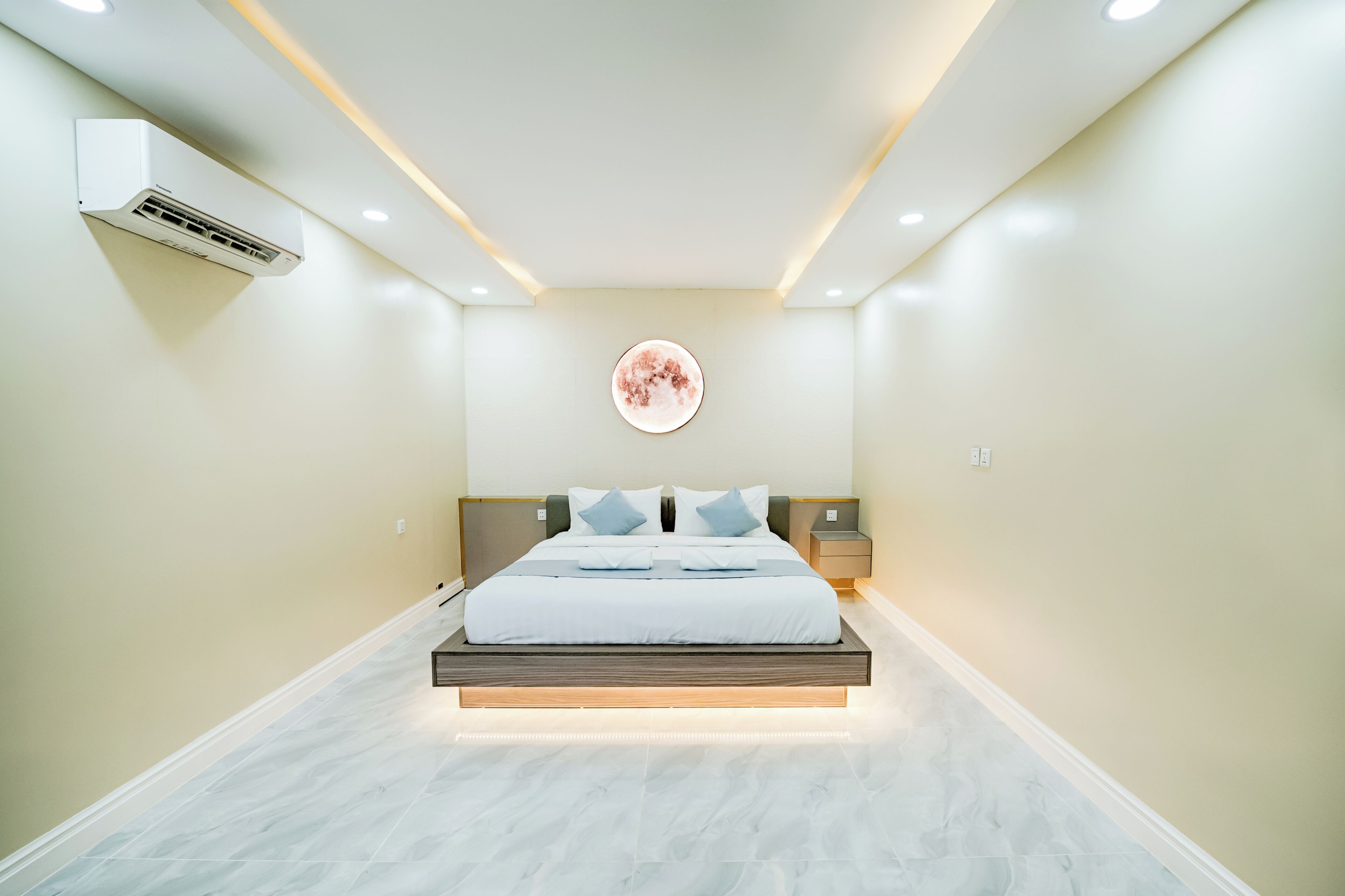 Ein modernes Schlafzimmer mit einem Bett, das auf einer Plattform zentriert ist, inklusive Einbauleuchten, einer wandmontierten Klimaanlage und einem runden Kunstwerk über dem Bett.