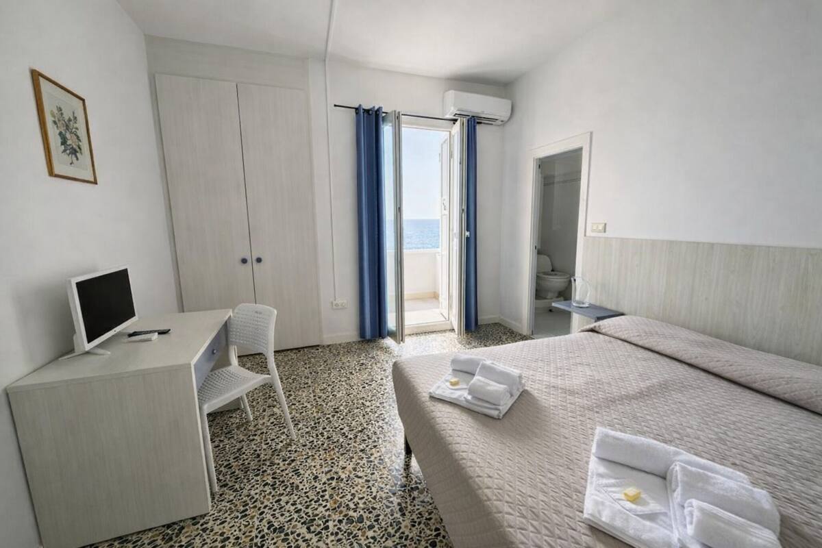A hotel room with a bed, a desk with a computer, a chair, a bathroom, and a view of the sea.