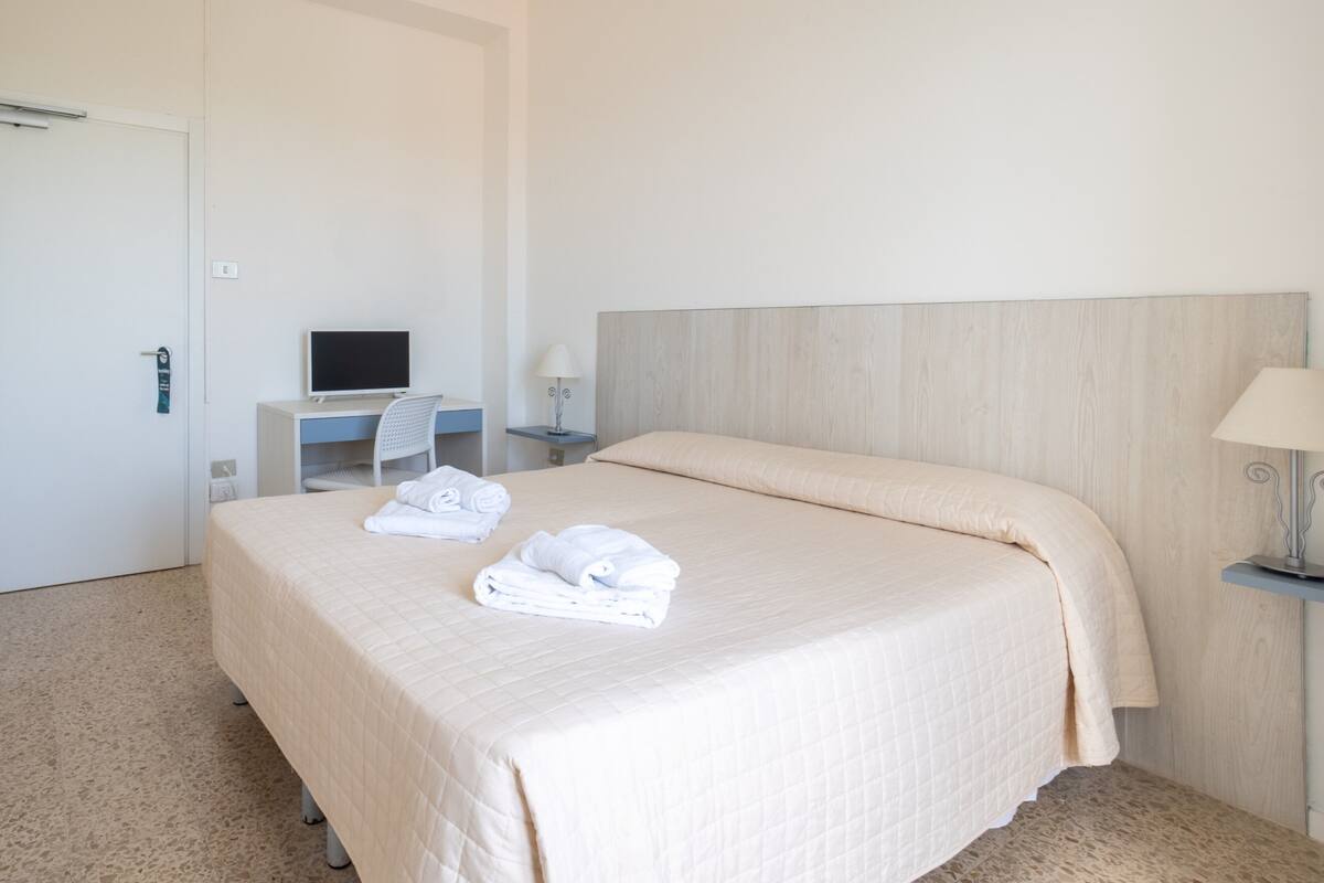 A hotel room with a bed, a desk with a computer, a chair, and two bedside lamps.