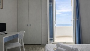 Ein Hotelzimmer mit Bett, Schreibtisch mit Computer, Stuhl und Blick aufs Meer durch eine offene Tür.