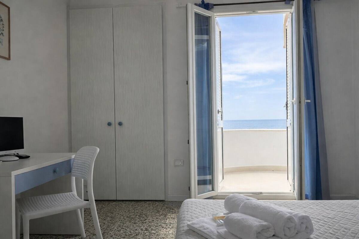 A hotel room with a bed, a desk with a computer, a chair, and a view of the sea through an open door.