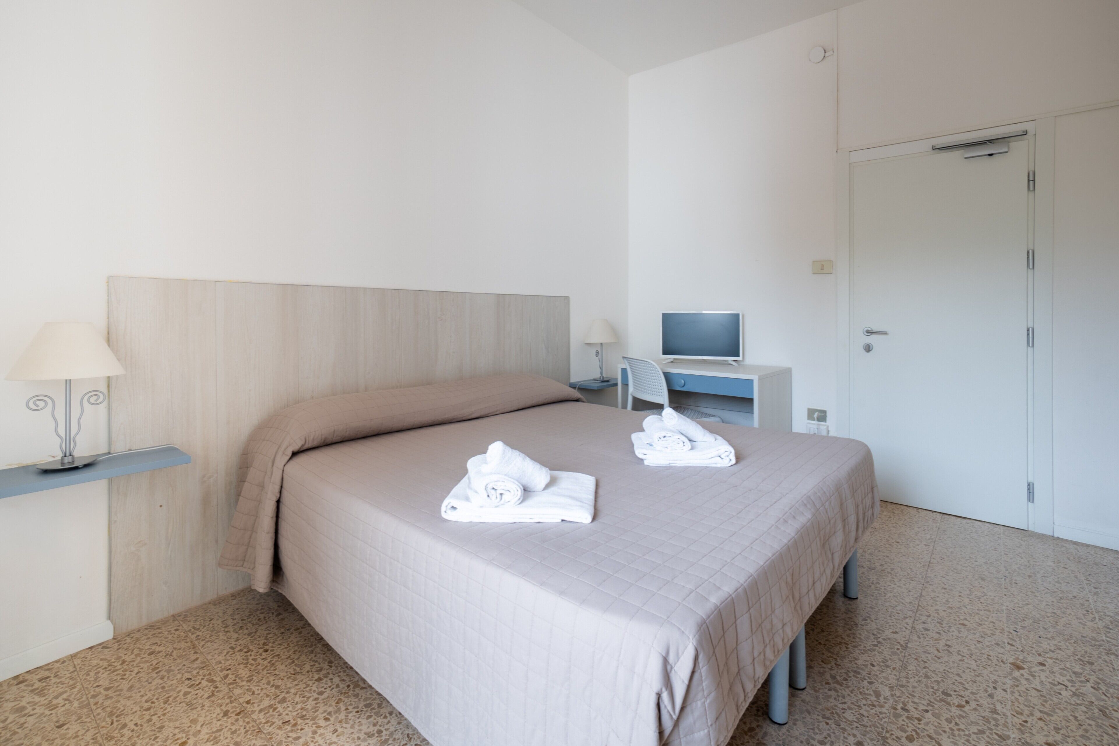 Ein Hotelzimmer mit einem Bett, einem Schreibtisch mit Computer, einer Lampe und einer Tür.