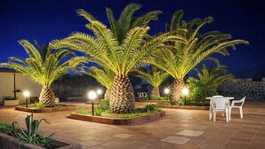 A patio area with palm trees, a table with chairs, and ambient lighting. - Hotiday Castelsardo Spiaggia (Lu Bagnu)