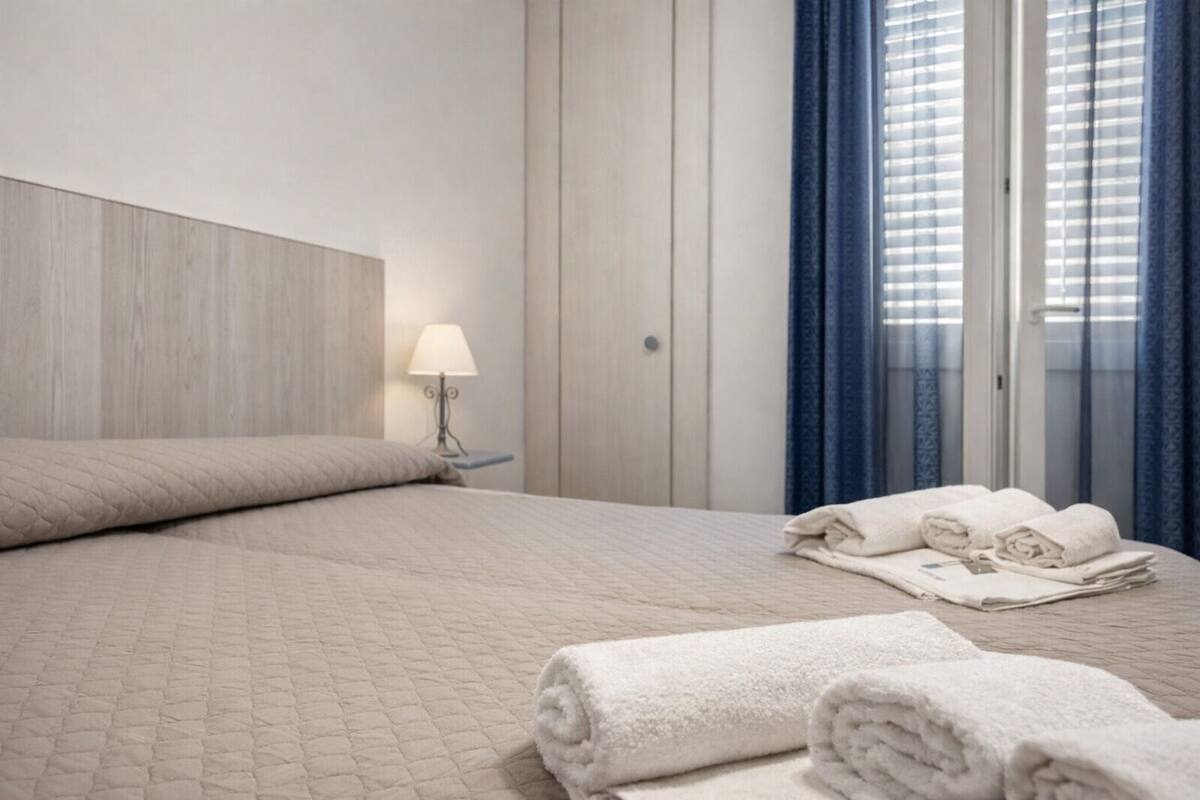 A hotel room with a bed, a nightstand with a lamp, a window with curtains, and a set of towels on the bed.