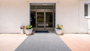 A building entrance with a glass door, potted plants, and a metal trash can. - Hotiday Castelsardo Spiaggia (Lu Bagnu)